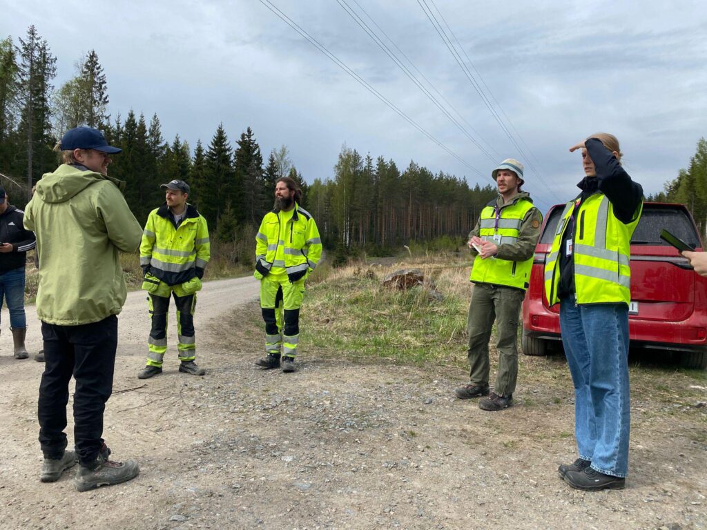 Henkilöitä huomioliivit ja -takit päällä maastossa, taustalla auto, hiekkatie ja sähkölinjoja